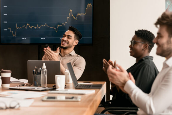 A diverse group of professionals clapping and smiling in a boardroom, with a financial chart displayed on a screen in the background, celebrating business success.